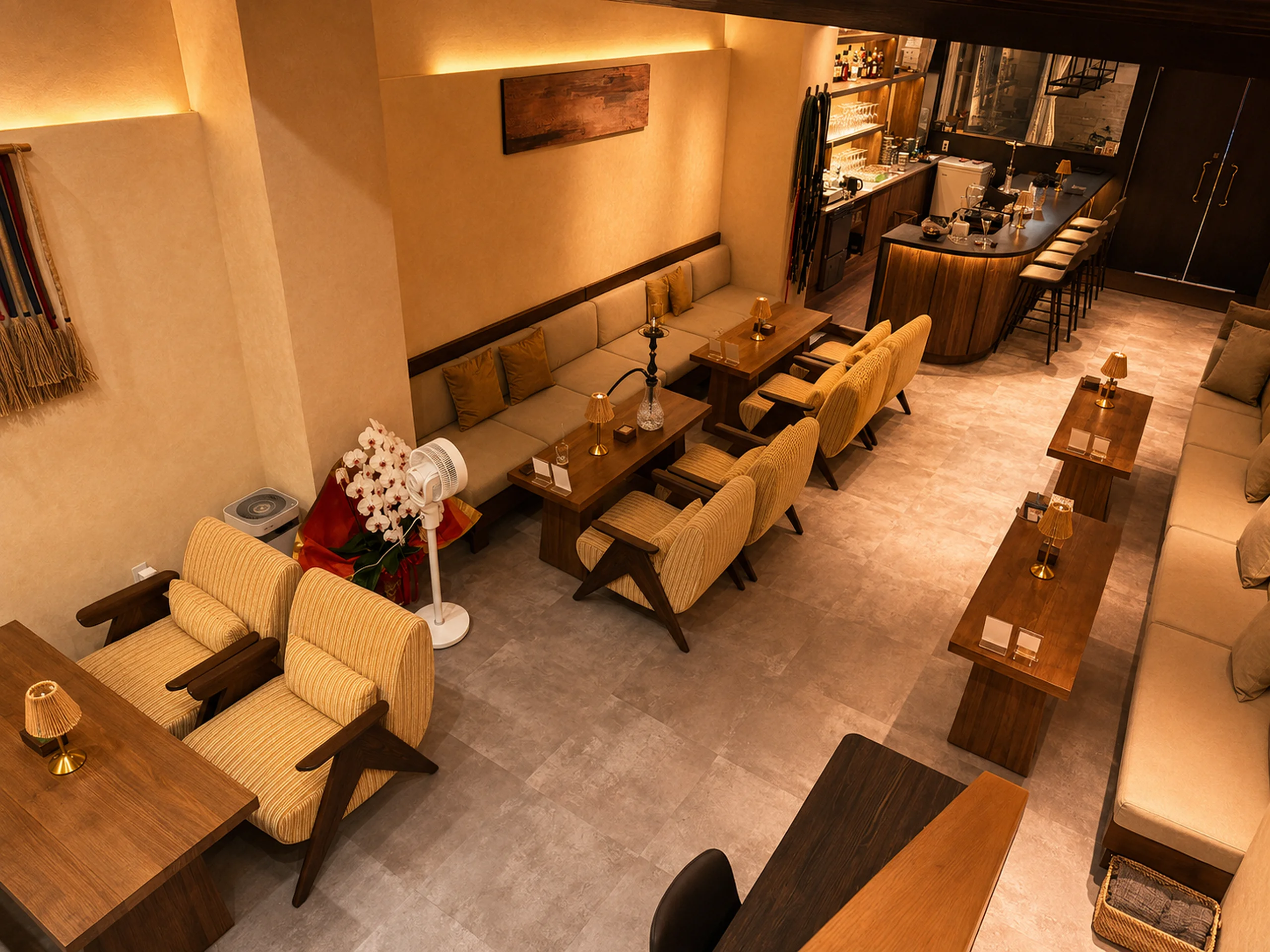 A wide view of the lounge — striped armchairs and low wooden tables with rattan lamps, banquettes along the wall, the bar counter at the back, a rust-toned panel and a woven wall hanging